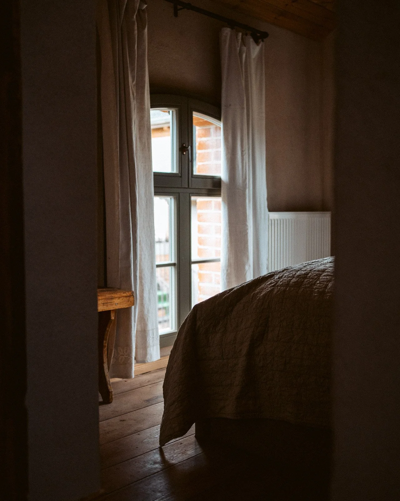 Ein Blick in ein Schlafzimmer mit einem Fenster, weichen weißen Vorhängen, einem Holzfußboden und einem Bett mit einer braunen Decke.