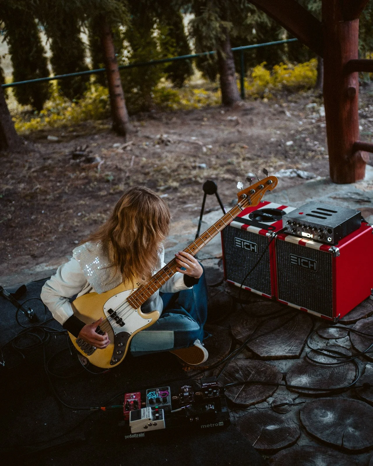 Eine Person mit langen, gewellten, braunen Haaren sitzt auf dem Boden und spielt ein Bassgitarrenspiel im Freien. Es gibt Verstärker und Pedale auf dem Boden neben ihr.