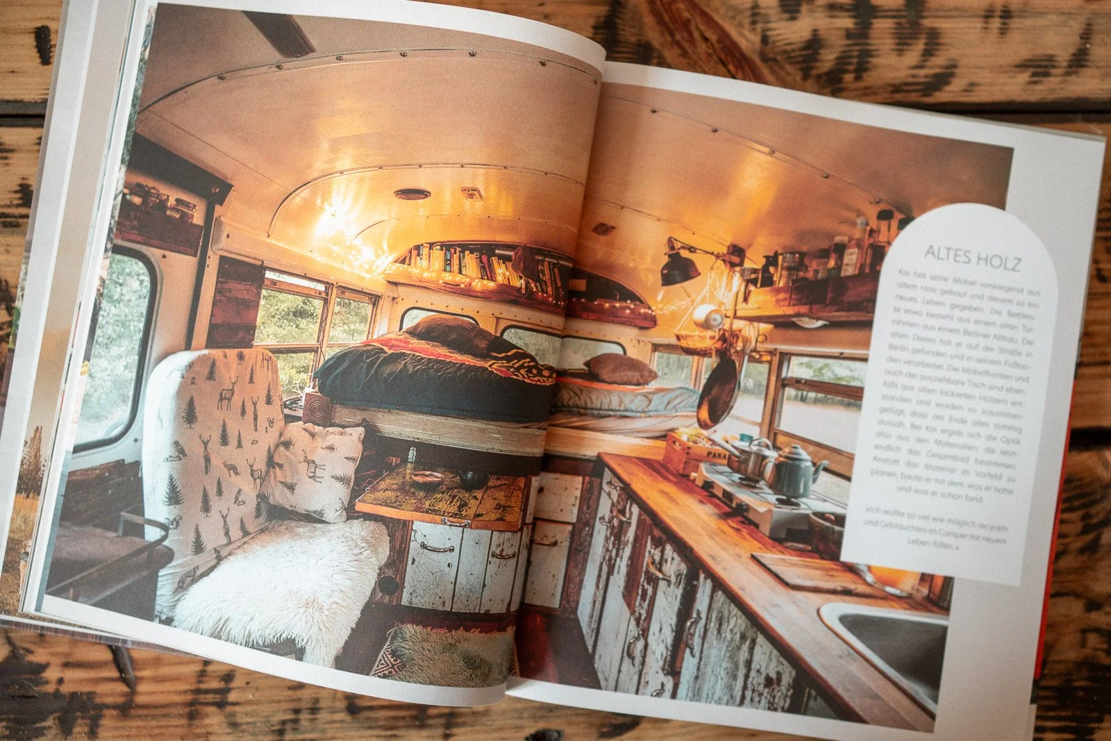 Innenansicht eines umgebauten Schulbusses mit Holzkomponenten, Bett, Sitzbereich, Bücherregal, Küchenzeile und Fenster