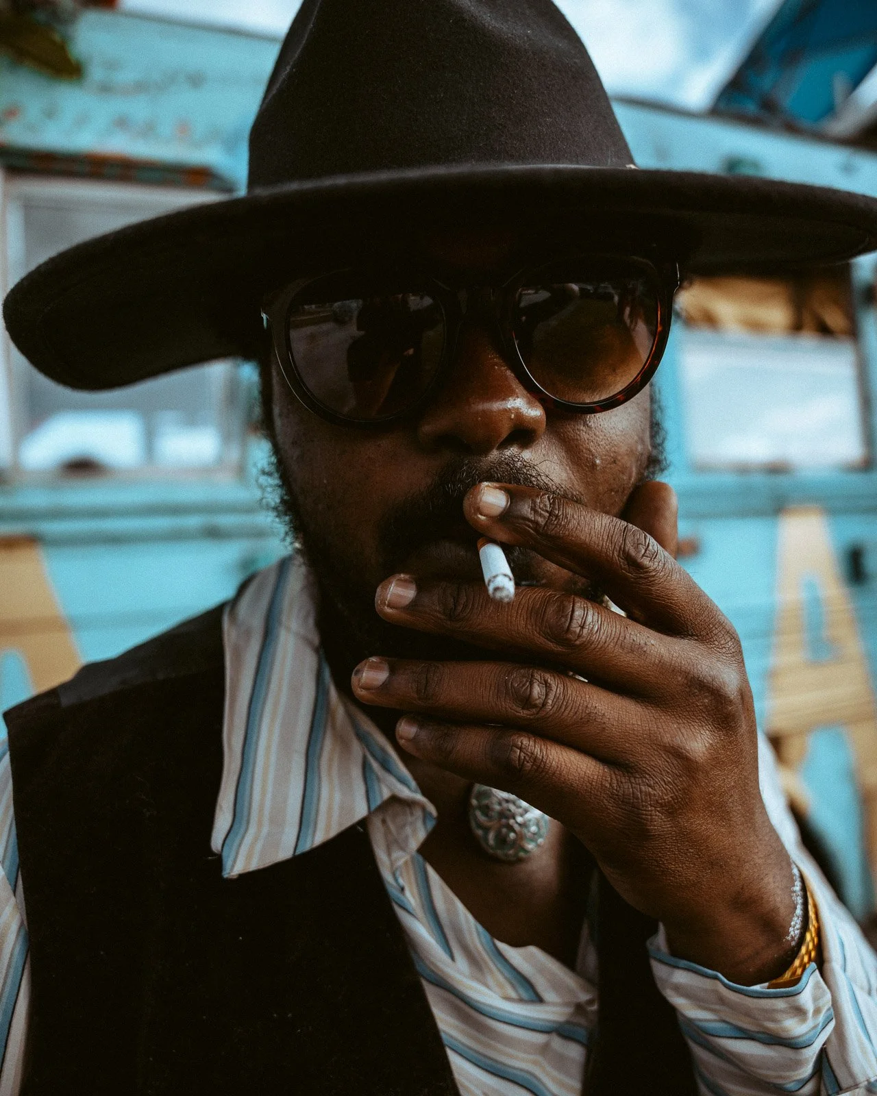Ein Mann trägt einen schwarzen Hut und dunkle Sonnenbrillen, raucht eine Zigarette, trägt ein gestreiftes Hemd und einen schwarzen Weste, und hat mehrere Schmuckstücke, darunter ein Halskette und ein Armband.