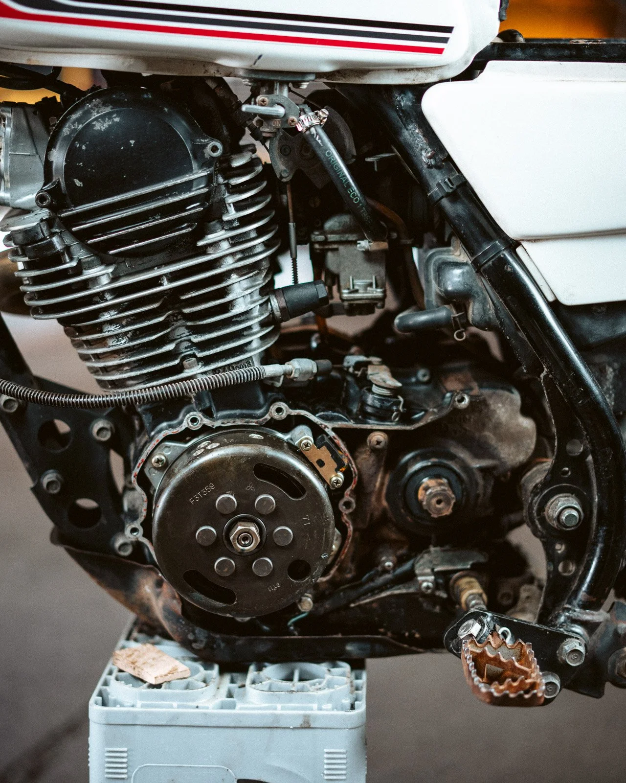 Nahaufnahme eines Motorradmotors, der teilweise zerlegt wurde, auf einer weißen Plastikbox, mit sichtbaren Schrauben, Riemen und Metallteilen.