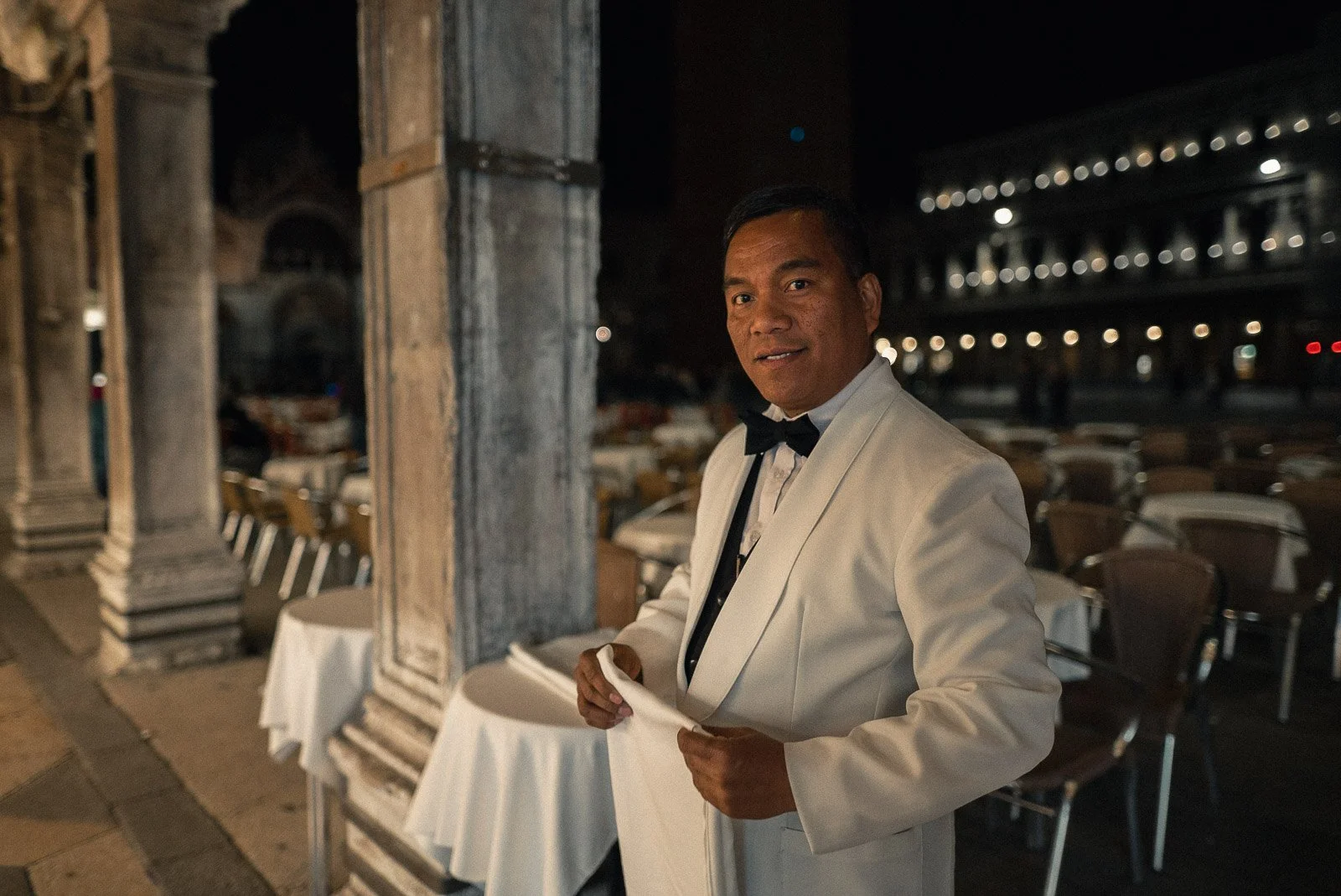 Ein Mann in einem weißen Anzug und Fliege steht in einem eleganten Restaurant bei Nacht, mit Tischen und Stühlen im Hintergrund.