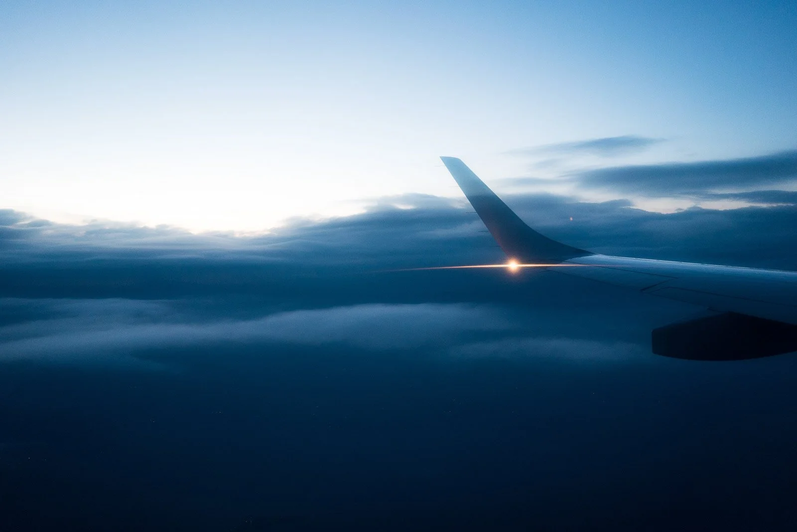Blick auf eine Flugzeugflügel im Flug bei Sonnenaufgang oder Sonnenuntergang, umgeben von Wolken und einem blauen Himmel.