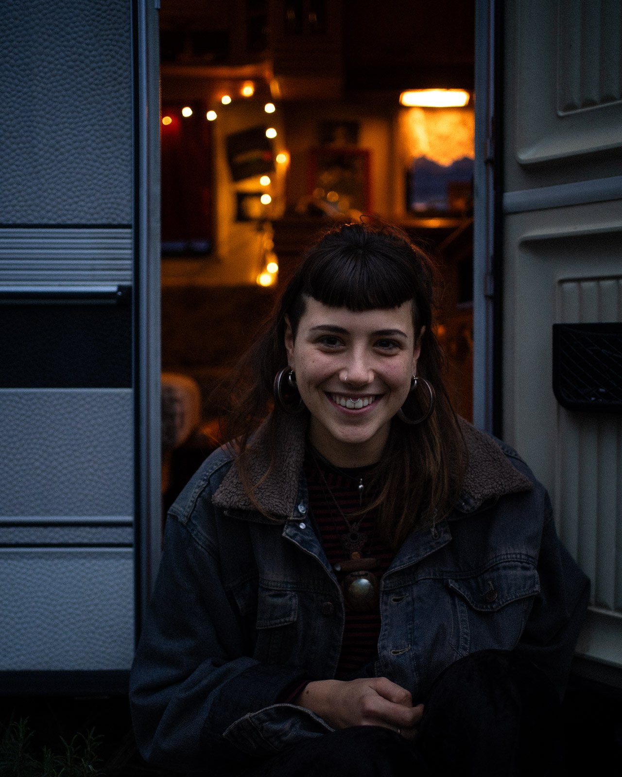 Junge Frau sitzt vor einem Wohnmobil, bei Dämmerung, lachend, mit dunklem Haar und Jeansjacke.