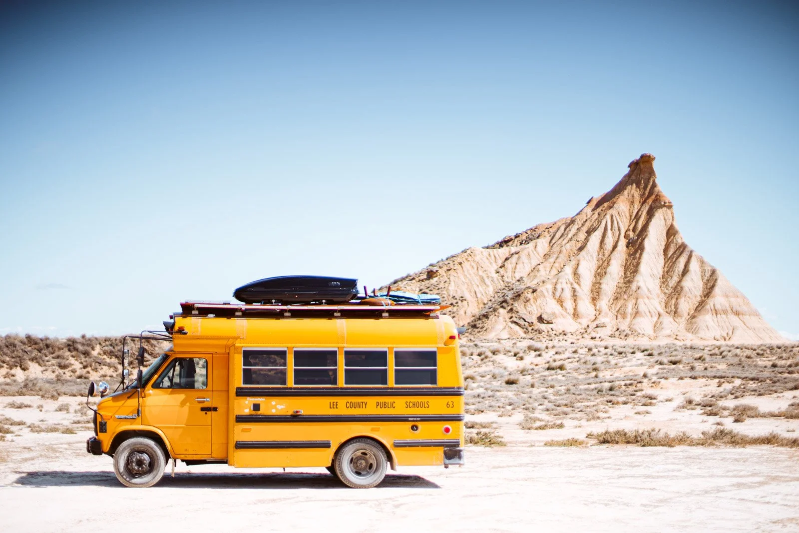 Eine gelbe Schulbus im Wüstengebiet mit einer markanten, bergähnlichen Felsformation im Hintergrund unter klarem blauen Himmel.