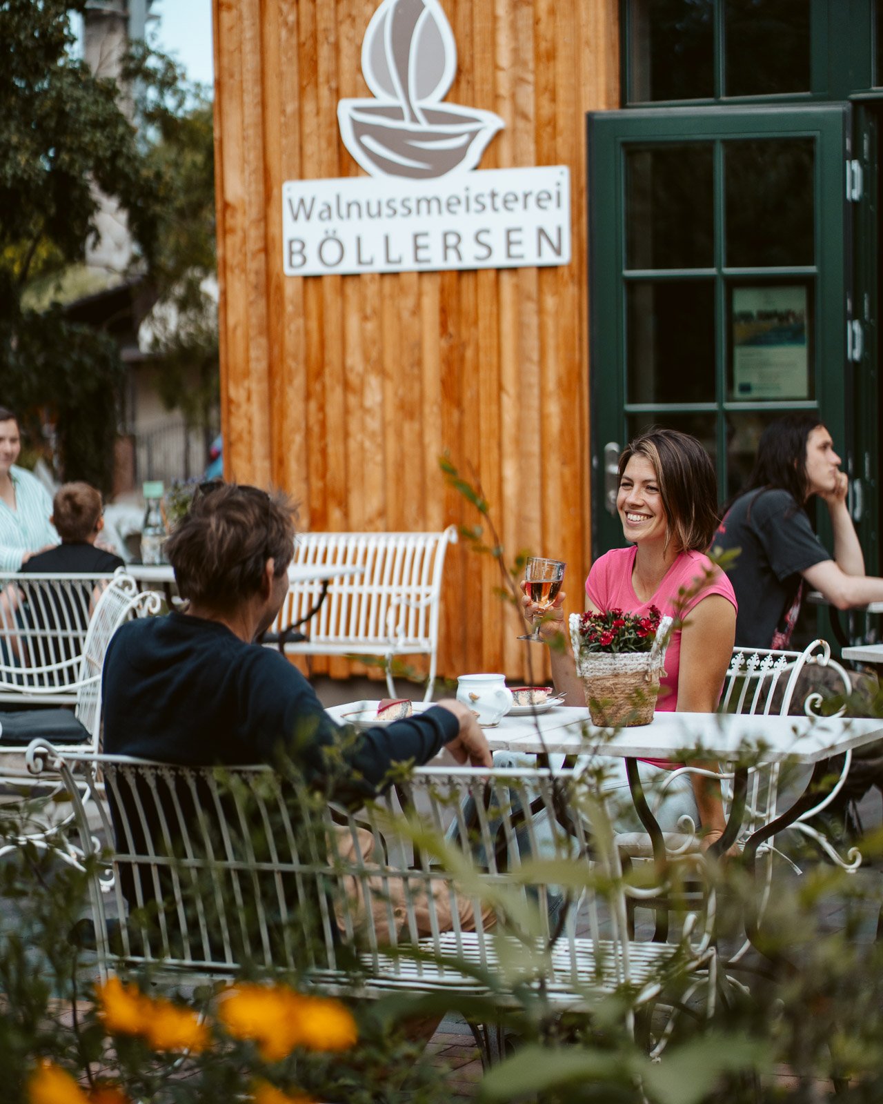 Frauen sitzen draußen an einem Tisch, trinken Wein und unterhalten sich vor einem Holzhaus mit einem Schild der Walnussmanufaktur BOLLERSEN.