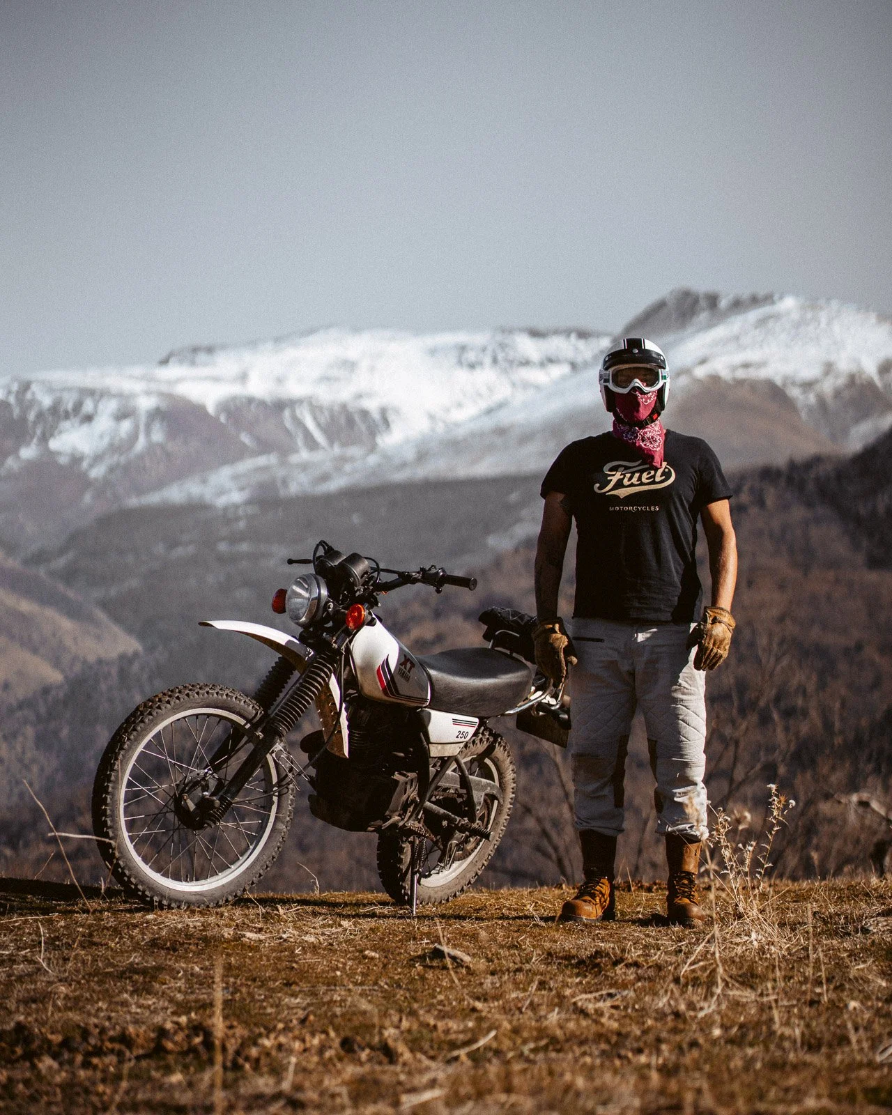 Ein Mann mit Helm, Schutzbrille, Mundschutz, Handschuhen, schwarzem T-Shirt, zerrissenen Hosen und grauen Stiefeln steht neben einem weißen Motorrad vor einer Bergkulisse mit schneebedeckten Gipfeln.