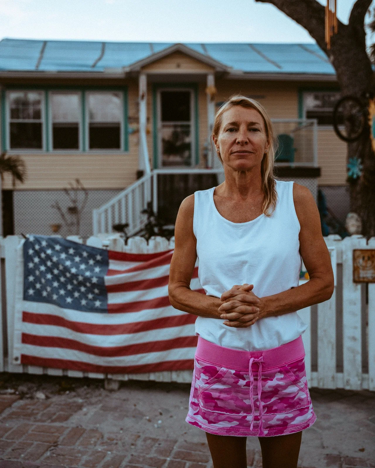Eine ältere Frau steht vor einer weißen Gartenzaun mit einer amerikanischen Flagge, im Hintergrund ein Haus mit blauen Fenstern und einer Veranda, bei Sonnenuntergang.