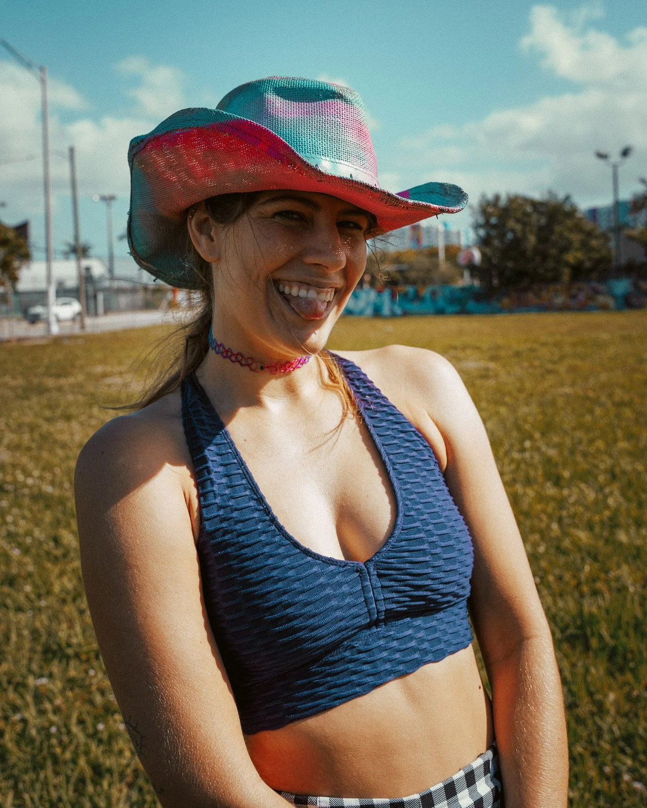 Lächelnde junge Frau mit bunter Cowboy-Hut im Freien, trägt ein blauen Sport-BH, trägt einen farbigen Choker, im Hintergrund eine Wiese und eine Stadt bei Sonnenschein.