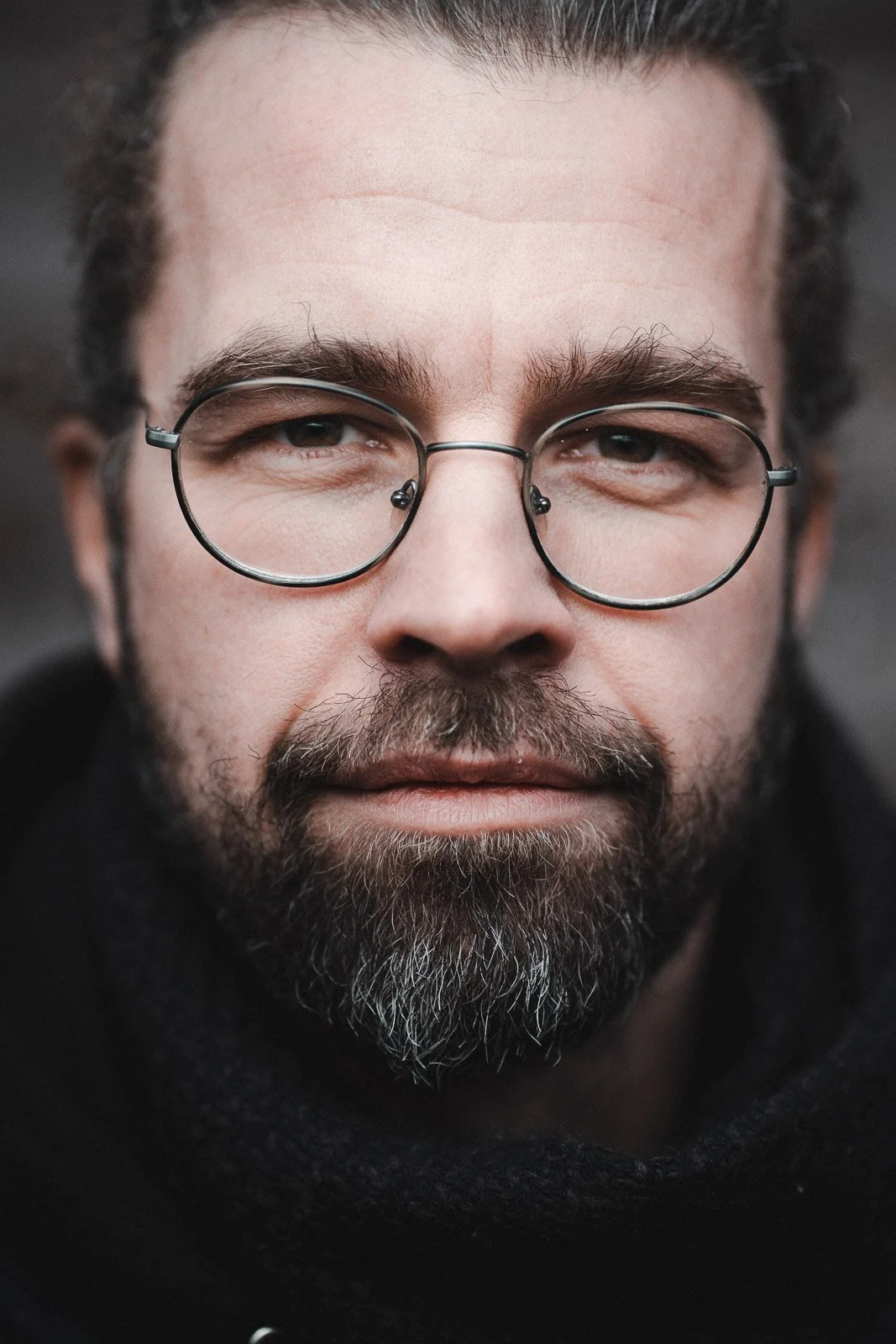 Nahaufnahme eines Mannes mit Bart, Brille und dunklem Hoodie, blickt direkt in die Kamera.