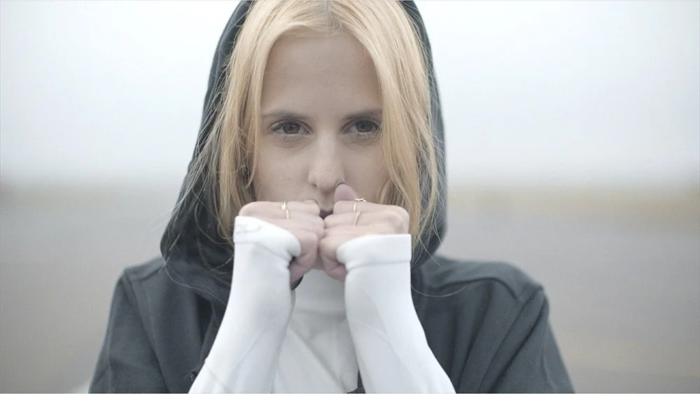 Junge Frau mit blondem Haar, die in eine Kamera schaut und ihre Hände vor dem Gesicht hält, trägt eine schwarze Jacke und weiße Ärmel.