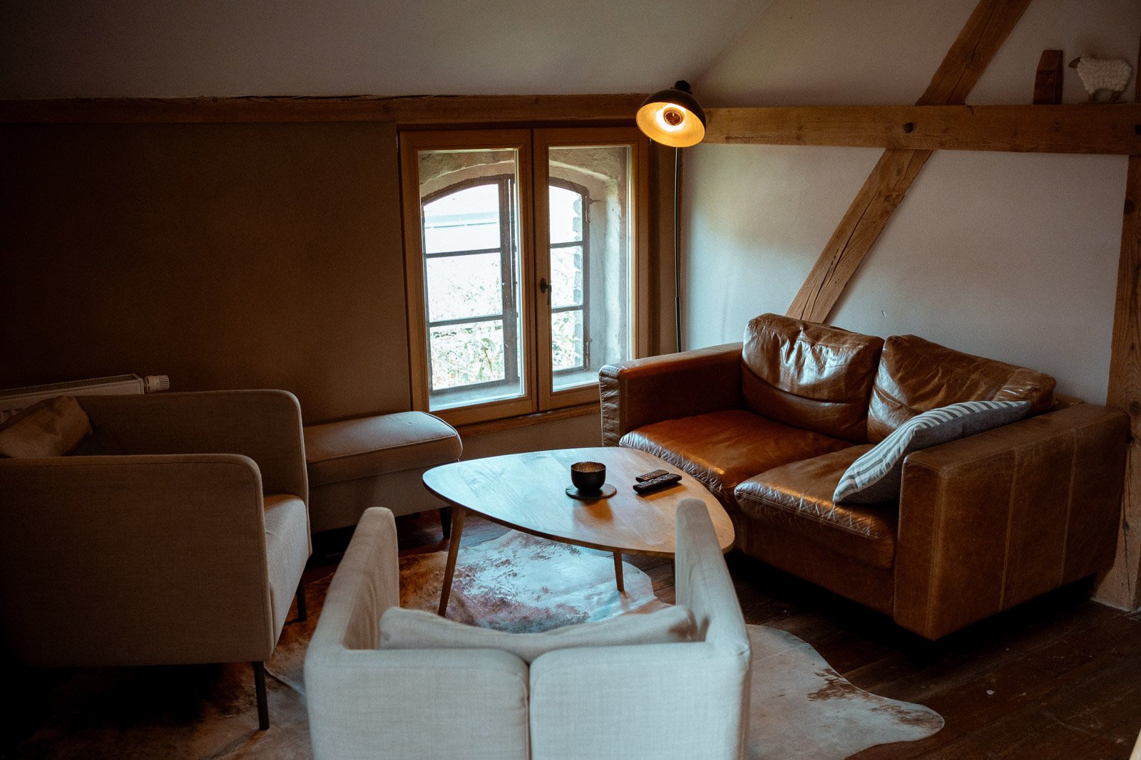 Gemütliches Wohnzimmer mit braunem Ledersofa, weißen Armchairs, Holztisch, Fenster mit Blick nach draußen, weiche Beleuchtung.