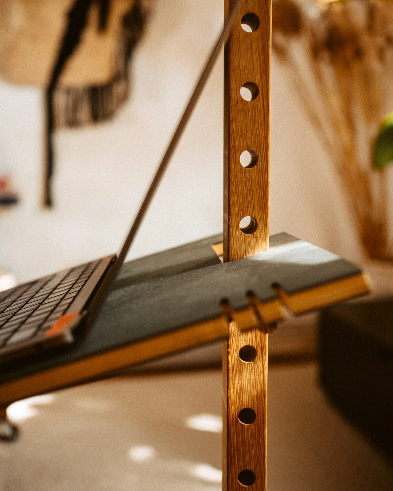 Nahaufnahme eines Holzregals mit runden Löchern, auf dem ein Laptop mit schwarzer Tastatur steht. Im Hintergrund sind Wanddekorationen sichtbar.