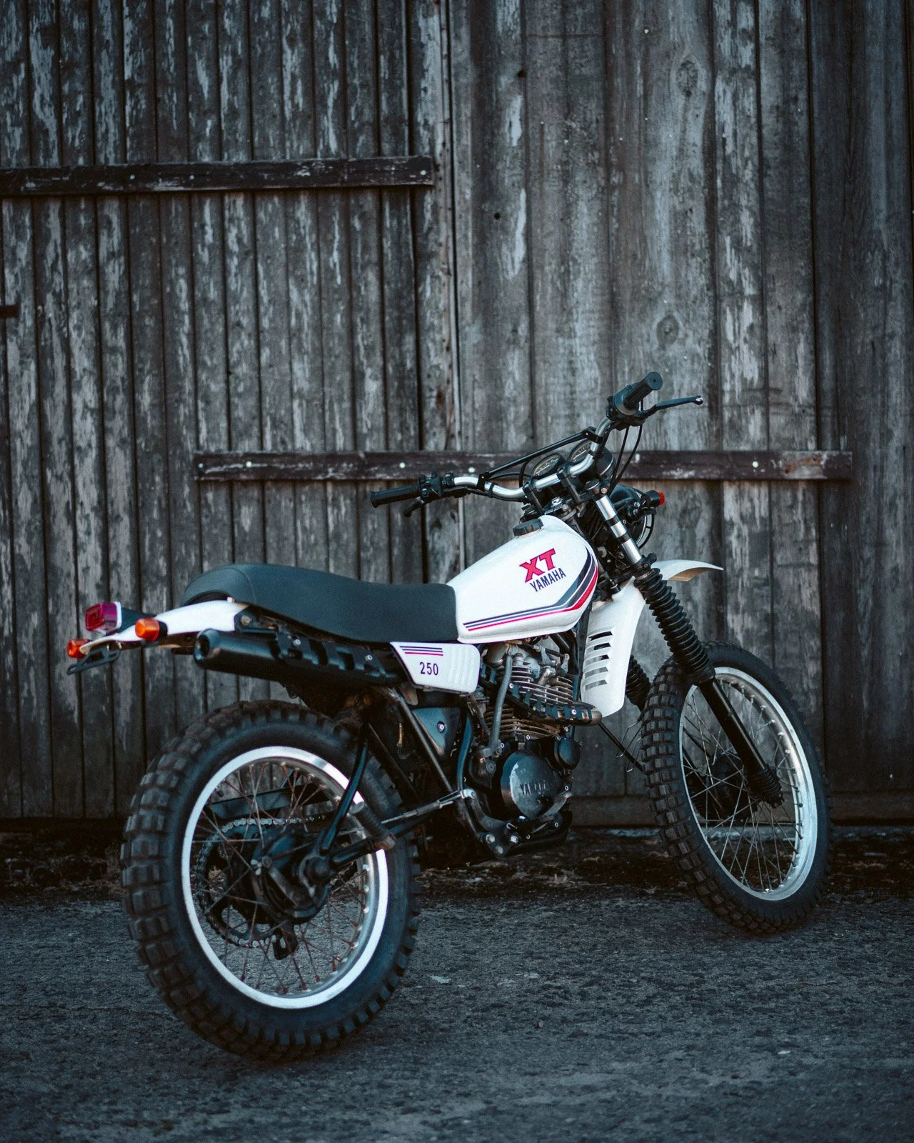 Ein weißes Yamaha XT Enduro-Motorrad mit schwarzen Reifen und knallhartem Gelände vor einer alten Holztür.