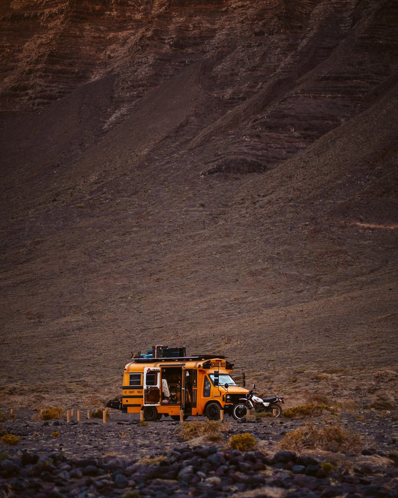Ein Schulbus und ein Motorrad stehen in einer einsamen, mondähnlichen Landschaft mit karger Vegetation und steilen Hügeln im Hintergrund.