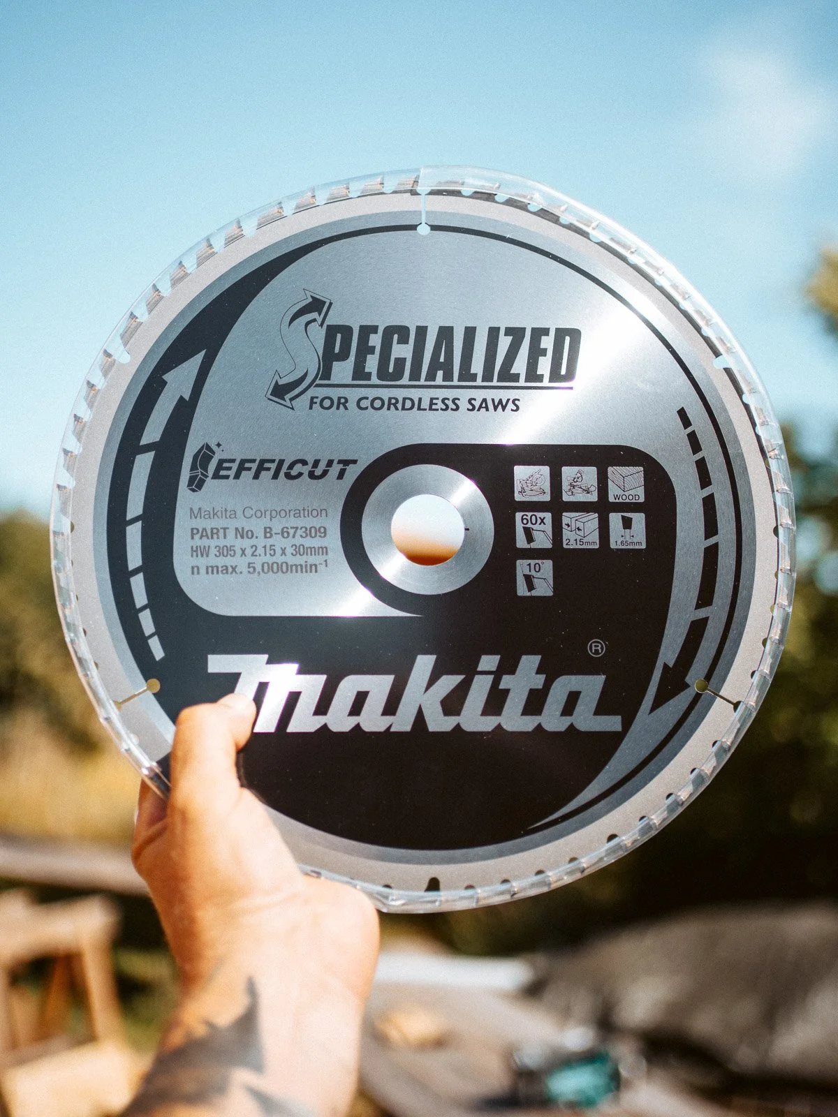 Nahaufnahme eines Kreissägeblatts mit Aufschrift 'Makita' und technischen Angaben, gehalten von einer Hand vor einem blauen Himmel im Freien.
