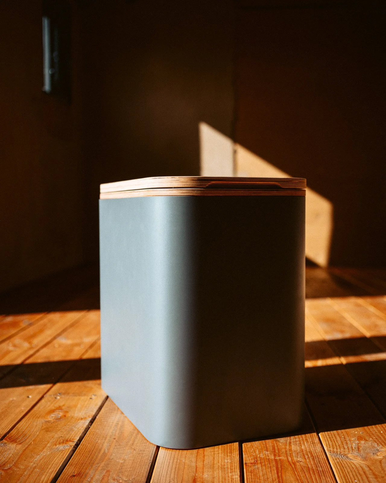 Ein dunkelgrauer, runder Holztruhe mit Holzdeckel steht auf einem Holzfußboden, beleuchtet durch Sonnenlicht.