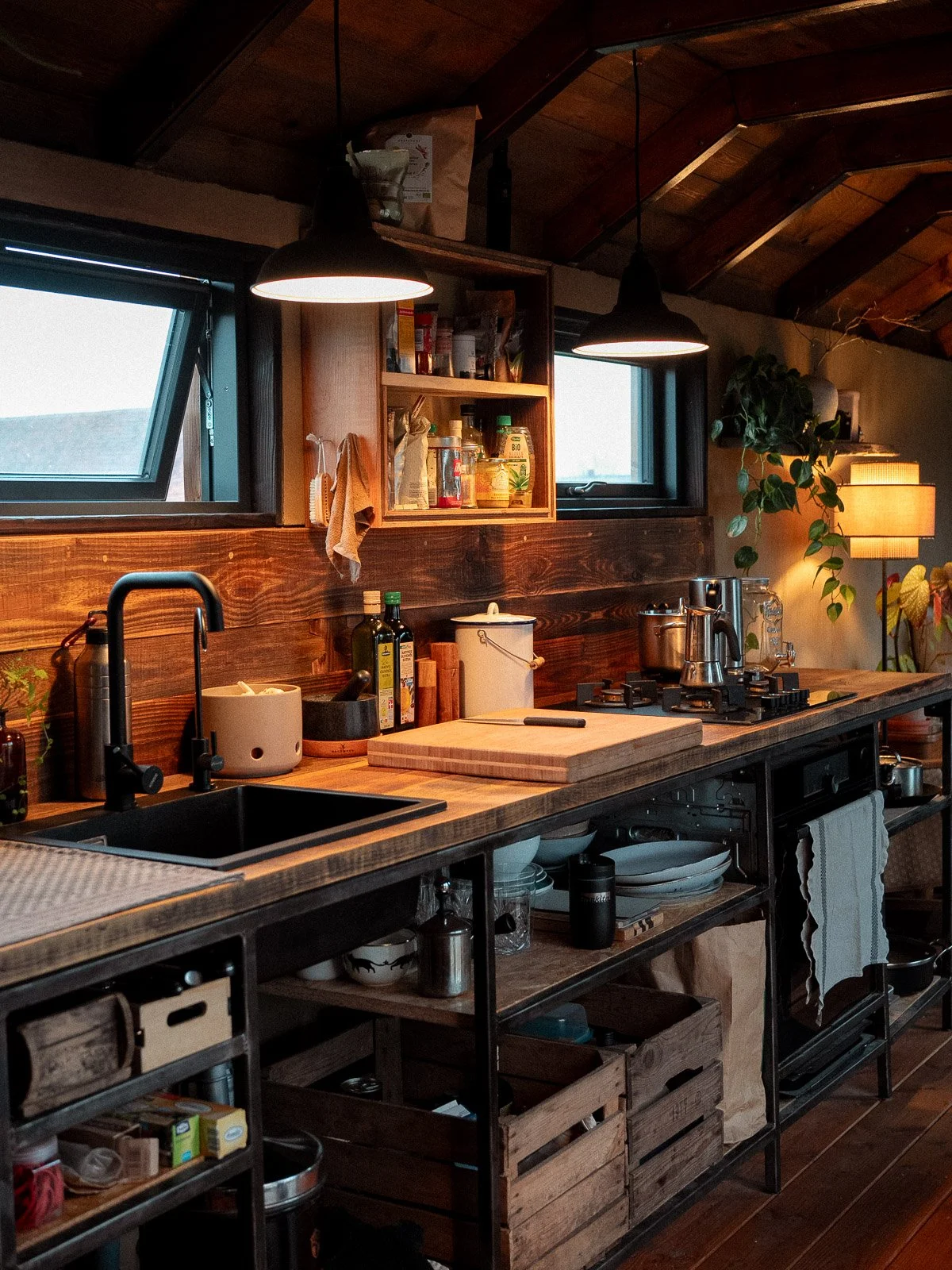 Küchenbereich mit Holzarbeitsplatte, Spüle, Kochfeld, offenen Regalen und Küchenutensilien, zwei kleine Fenster und eine Stehlampe im Hintergrund.