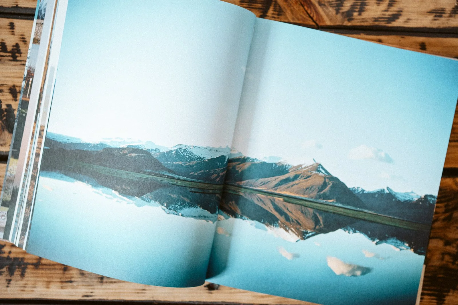 Buch mit einer Fotografie von Bergen und Wasser, die im Wasser gespiegelt sind, liegt auf einem hölzernen Tisch.