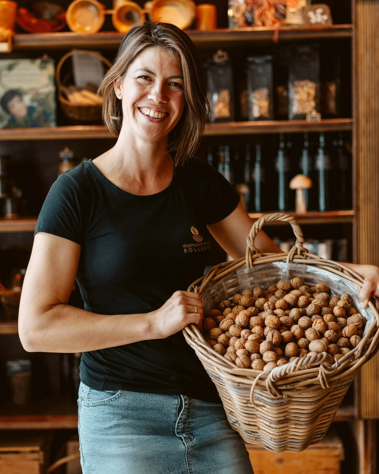 Eine Frau mit braunen, schulterlangen Haaren lächelt in die Kamera, sie hält einen Korb voller Walnüsse in einem Laden, im Hintergrund sind Holzbretter und verpackte Produkte sichtbar.