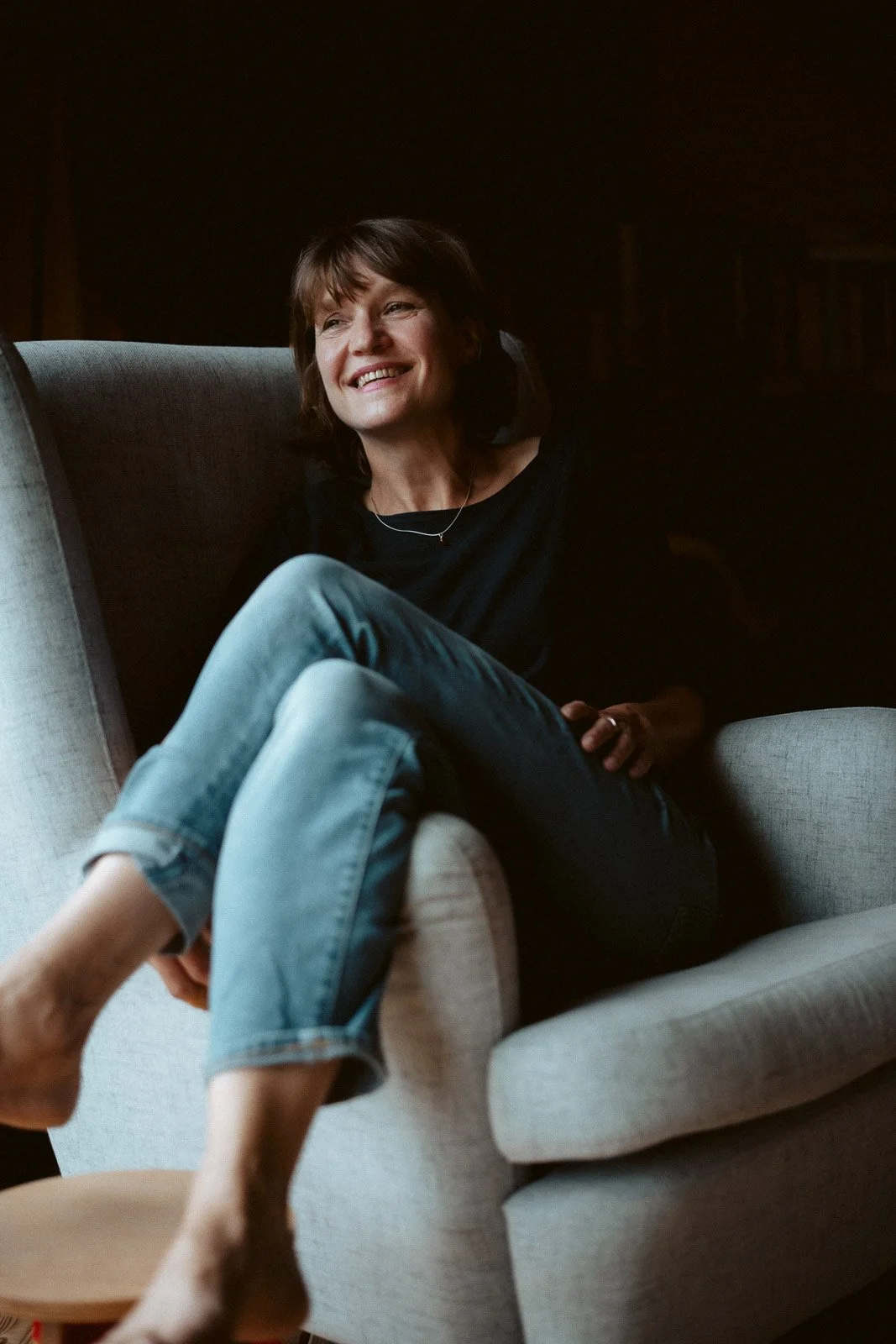 Frau entspannt in einem bequemen Sessel, lächelnd, in einem dunklen Raum, casual Kleidung, Jeans, kurzer Haarstil.