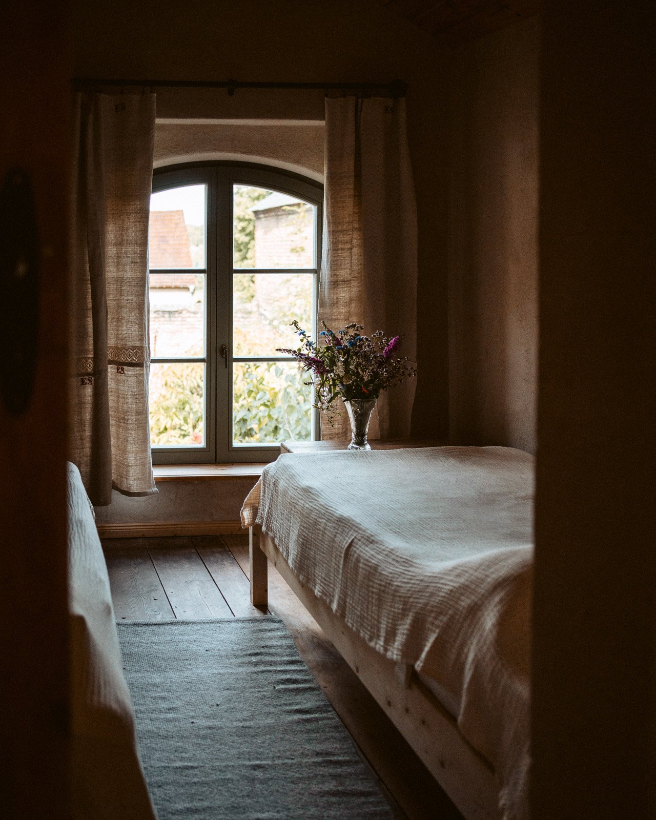 Ein Schlafzimmer mit einem Holzboden, einem Fenster mit weißen Vorhängen und einem Bett mit weißer Bettwäsche. Auf dem Nachttisch steht eine Vase mit bunten Blumen, die nach draußen blicken.