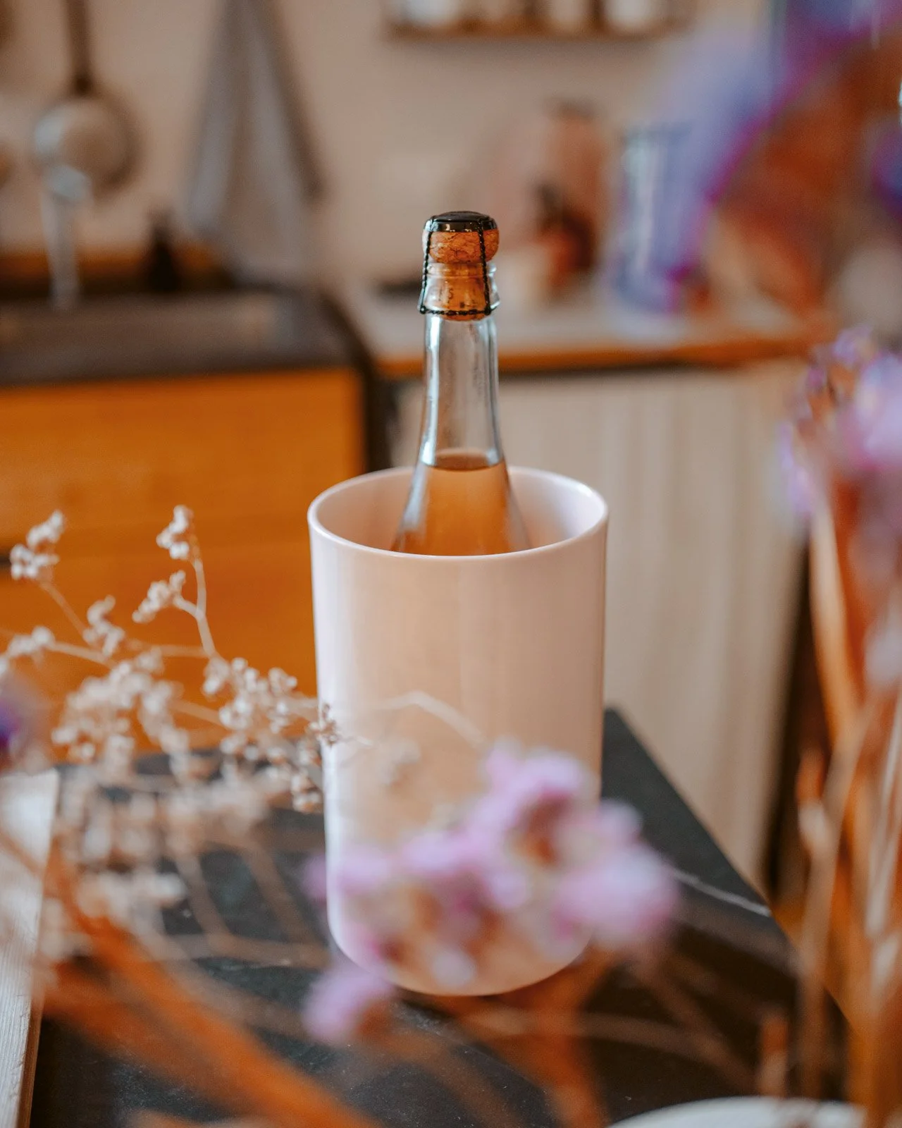 Eine leere Flasche steht in einem weißen Becher auf einem Tisch, umgeben von einigen Blumen, im Hintergrund eine Küche oder ein Esszimmer.