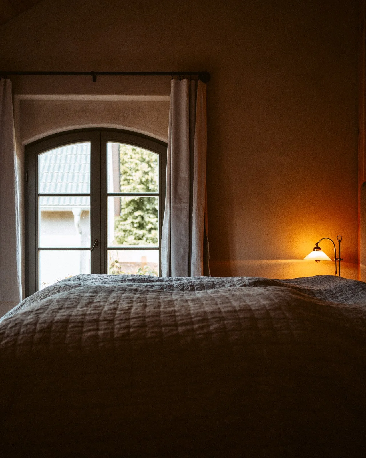 Ein Schlafzimmer mit einem Bett, das zu einem Fenster zeigt, das Vorhänge hat. Es ist abends, eine Lampe an der Wand ist eingeschaltet und strahlt warmes Licht aus.