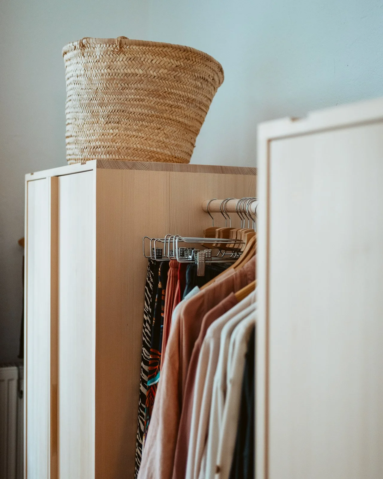 Ein Kleiderschrank mit hängender Kleidung und einem großen von Rattan geflochtenen Korb oben darauf.