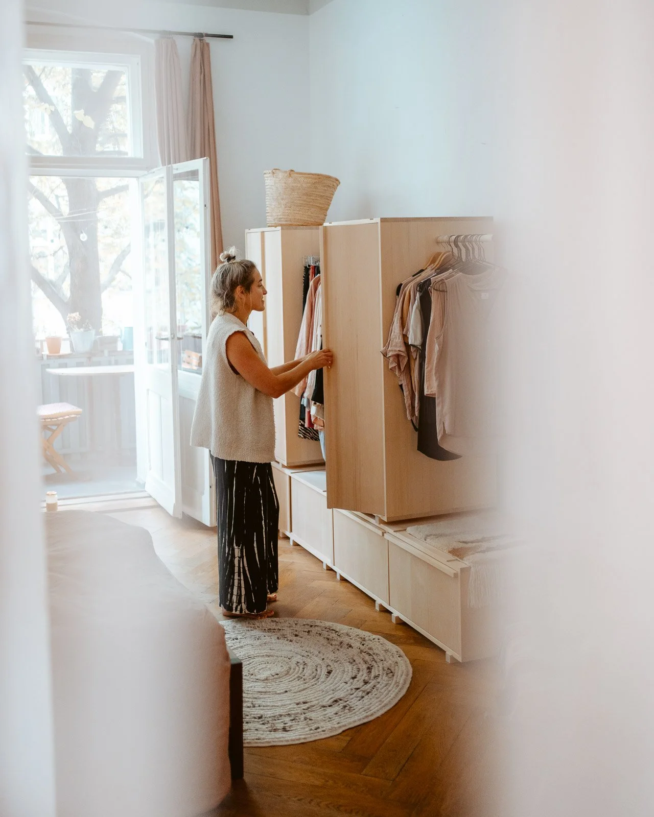 Eine Frau öffnet einen Kleiderschrank in einem hellen, gemütlichen Wohnzimmer mit Holzfußboden, Blick auf eine sonnige Terrasse.