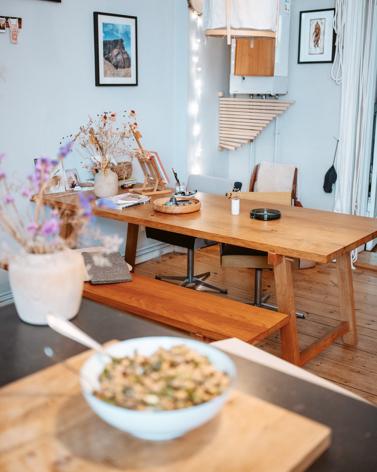 Ein Esszimmer mit einem Holztisch, bunten Blumen in einer Vase, mehreren Stühlen, Wandbildern und einer Thorwhelm-Lampe über dem Tisch. Im Vordergrund ist eine Schüssel mit Essen auf einem Schwarzen und den Holztisch zu sehen.