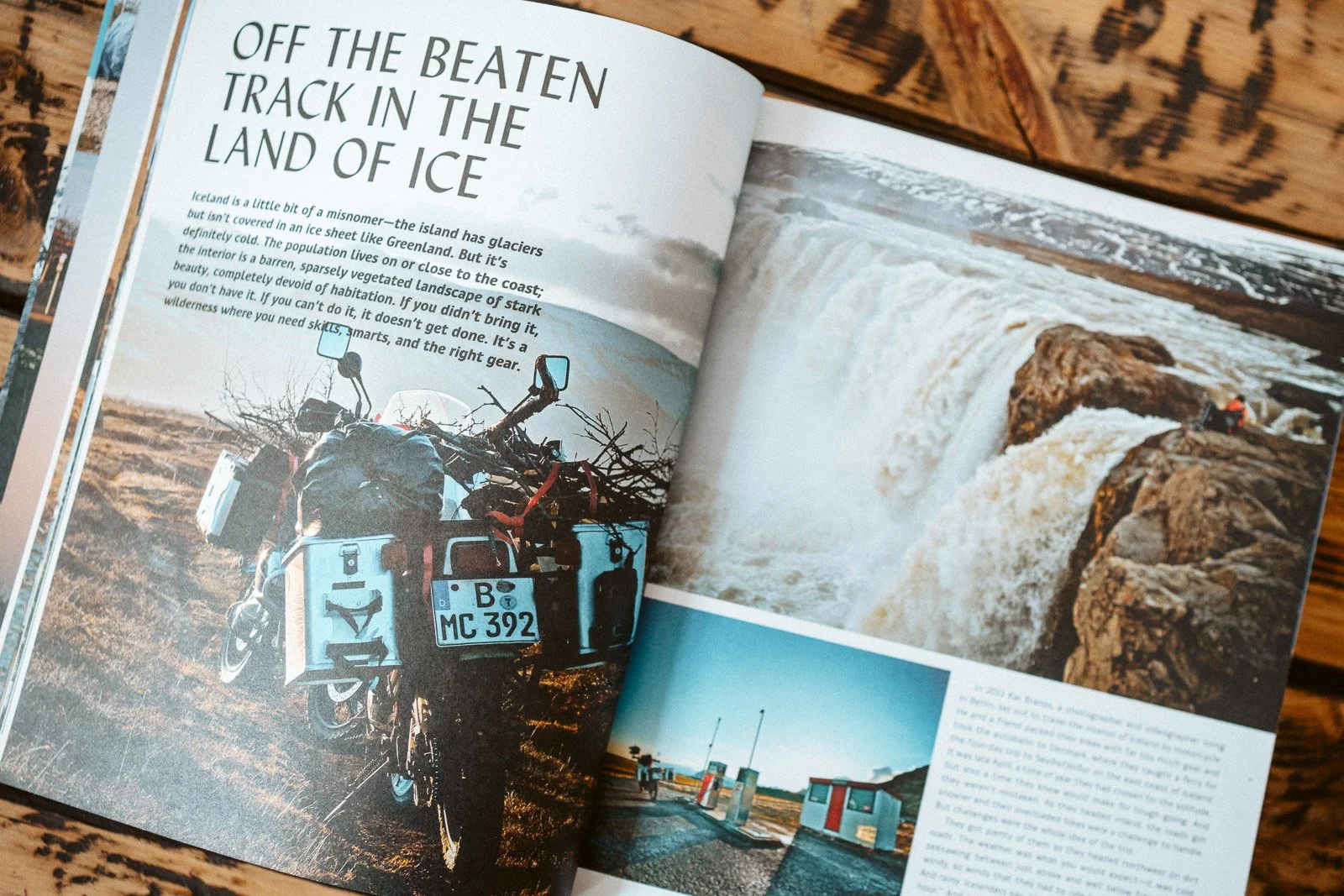 Ein offener Reiseführer auf einem Holzoberfläche zeigt eine Seite mit einem Motorrad mit Gepäck und einem deutschen Nummernschild, dazu ein Bild eines Wasserfalls und ein kleiner Fotocollage von einer Tankstelle und einem Gebäude an einem See.