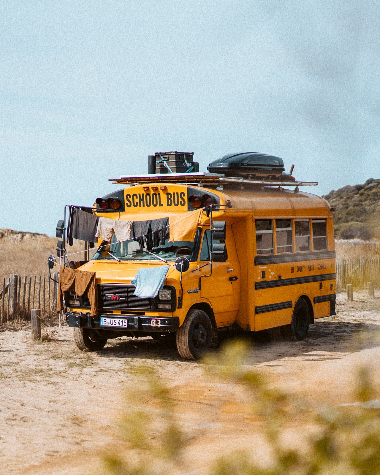 Ein gelber Schulbus steht am Strand, mit Wäsche zum Trocknen an den Fensterscheiben. Im Hintergrund sind Düne, Gras und Himmel sichtbar.