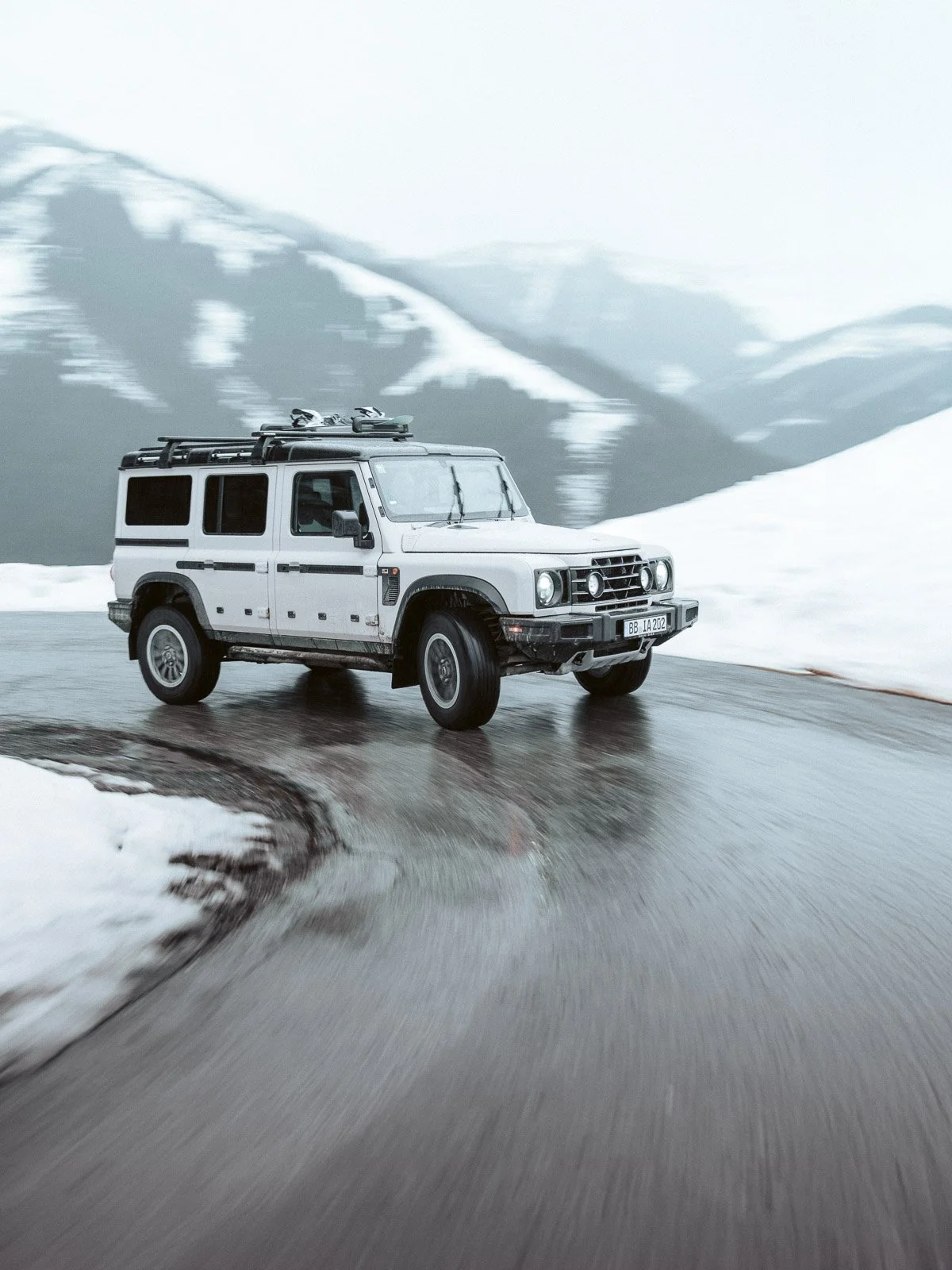 Ein weißer SUV fährt auf einer nassen, kurvigen winterlichen Straße durch schneebedeckte Berge