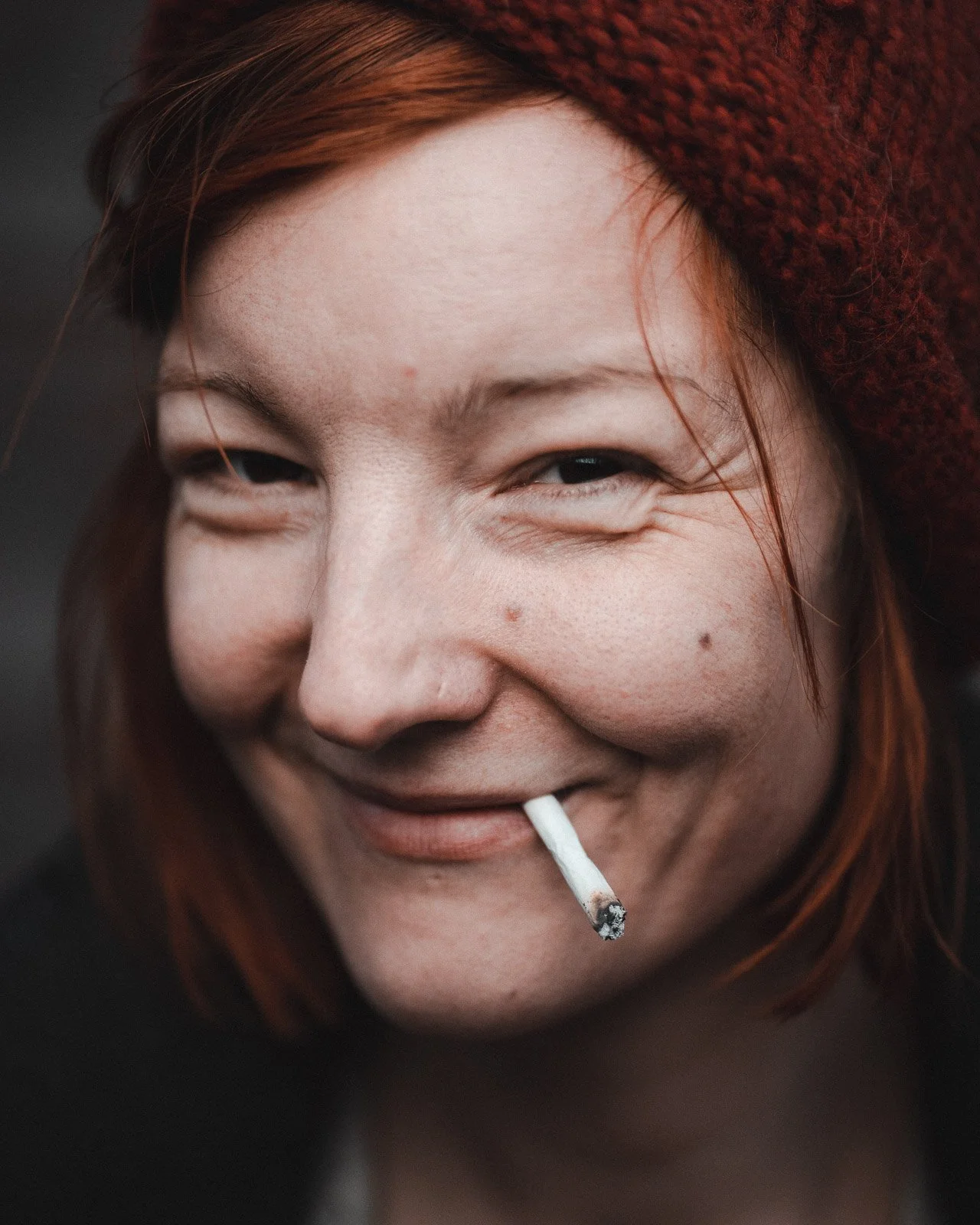 Nahaufnahme eines lachenden Frauenkopfes mit Zigarette im Mund, trägt eine rote Mütze und hat braune Haare.