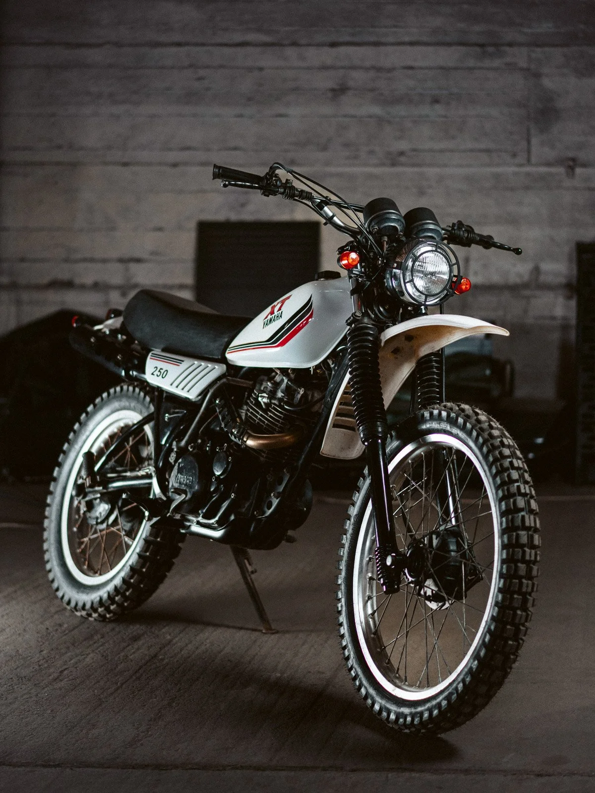 Ein Vintage-Motorrad in Weiß mit schwarzen und roten Akzenten, steht in einem dunklen Raum, mit sichtbaren Reifen, Scheinwerfer und Lenker.