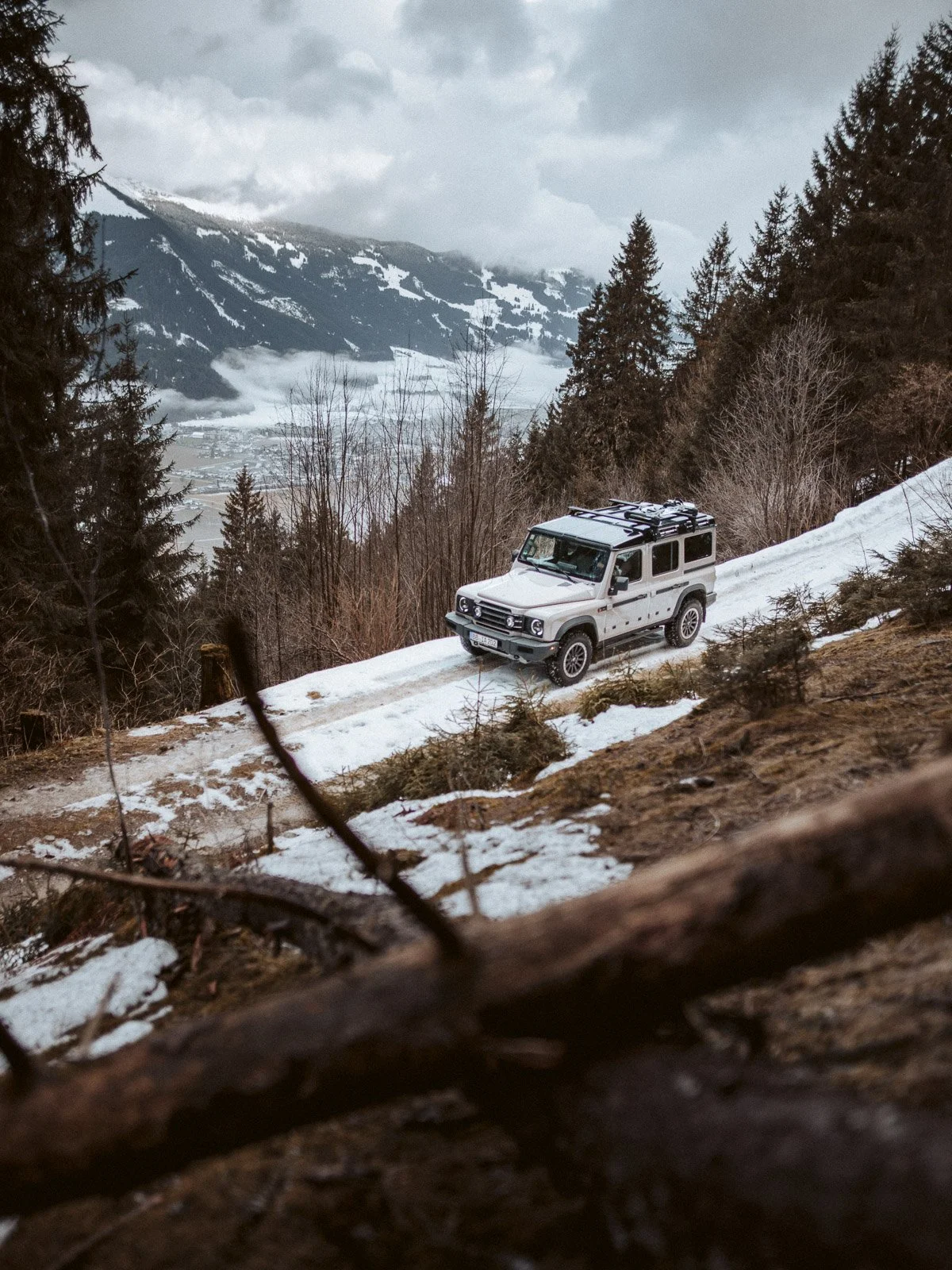 Ein weißer Geländewagen fährt auf einer schneebedeckten Straße in den Bergen, umgeben von Tannenbäumen und einer bewölkten Wetterkulisse.