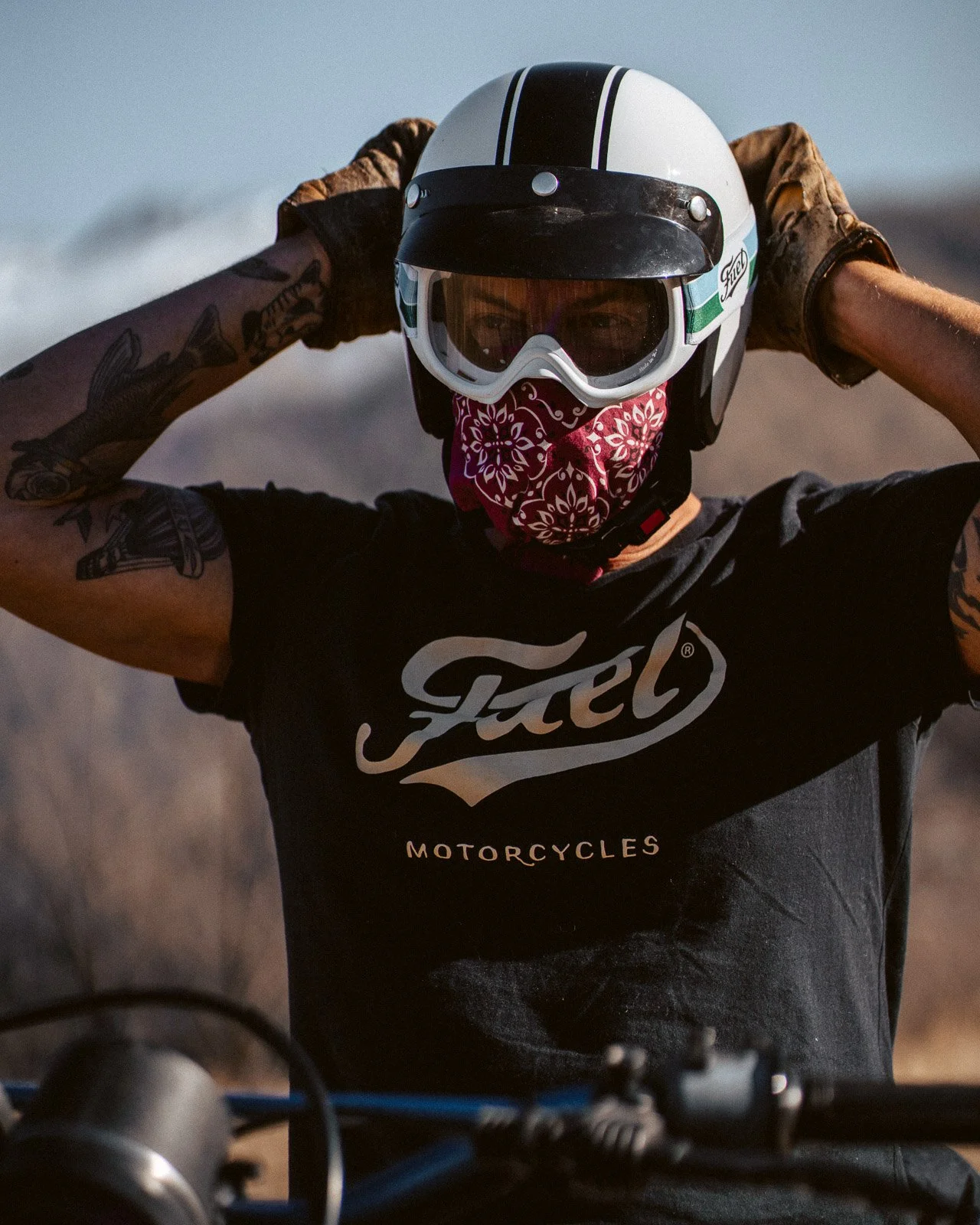 Motorradfahrer mit Helm, Sonnenbrille, Bandana und Tattoos, an einem Motorrad im Freien.