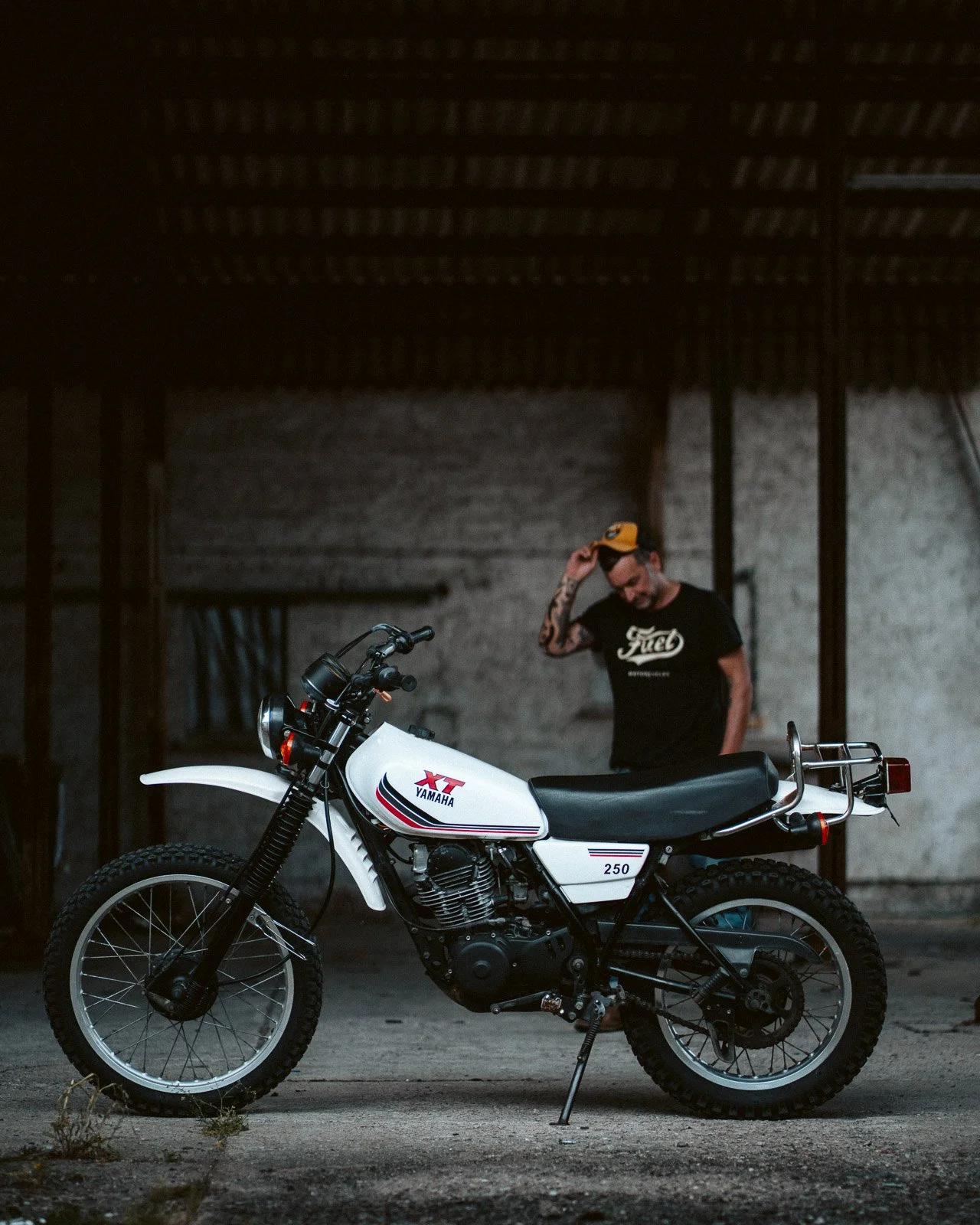 Ein weißes Yamaha XT 250 Enduro-Motorrad steht auf einem Betonboden in einem dunklen, industrialisierten Raum. Im Hintergrund ist ein Mann mit Tattoo, schwarz-weißes T-Shirt und einer gelben Motorradkappe, der nachdenklich schaut und die Kappe mit de