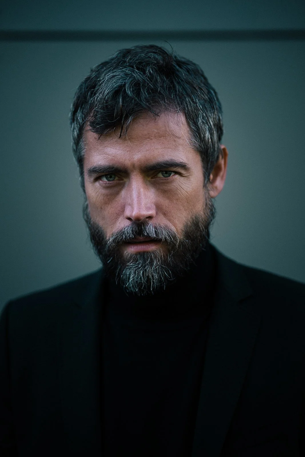 Porträt eines Mannes mit dunklem Haar und Bart, schwarzem T-Shirt und dunklem Hintergrund, ernsthafter Gesichtsausdruck.