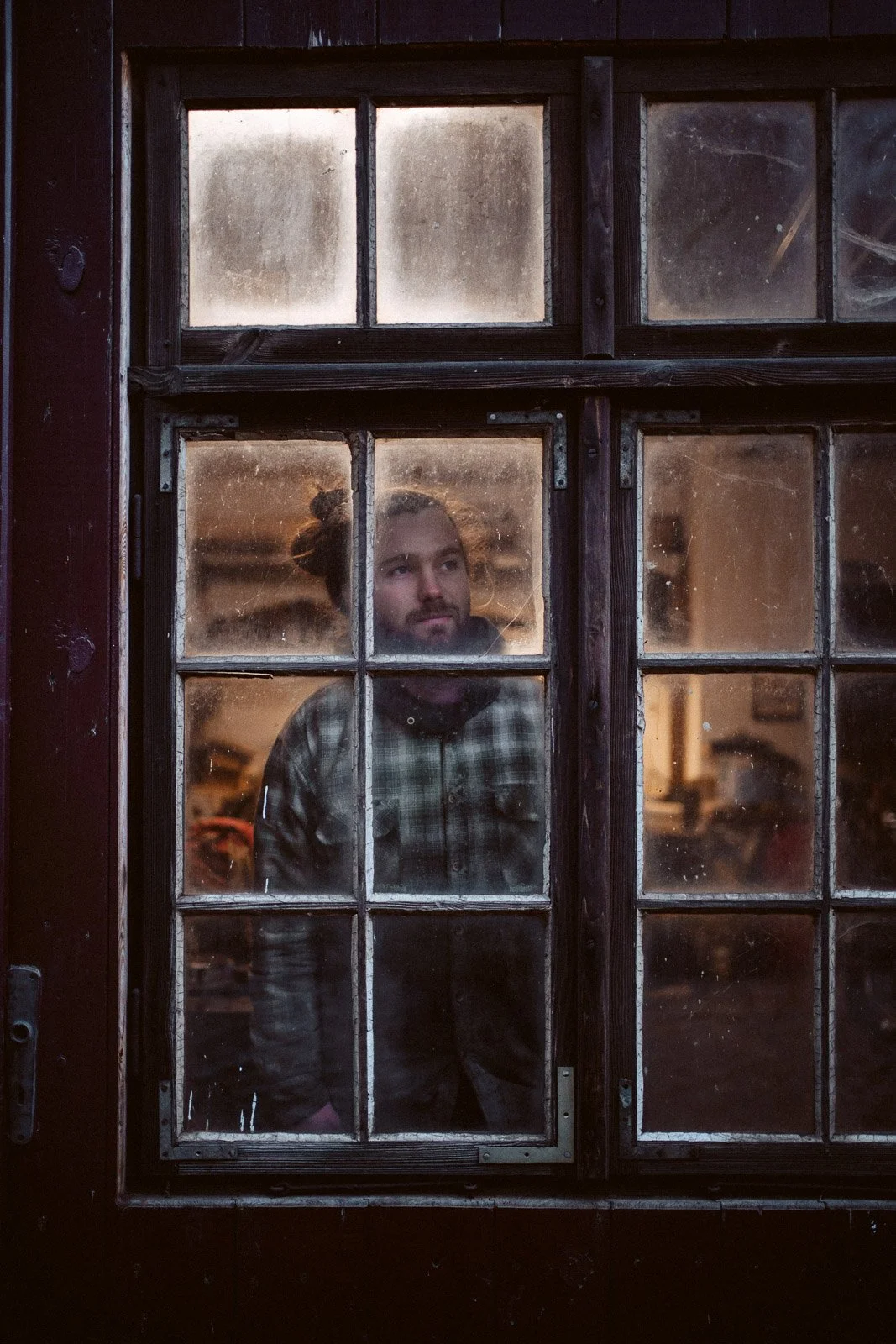 Ein Mann mit lockigem Haar, im Inneren eines Hauses, schaut durch ein altes, schmutziges Fenster. Es ist abends oder nachts, Licht ist im Raum.