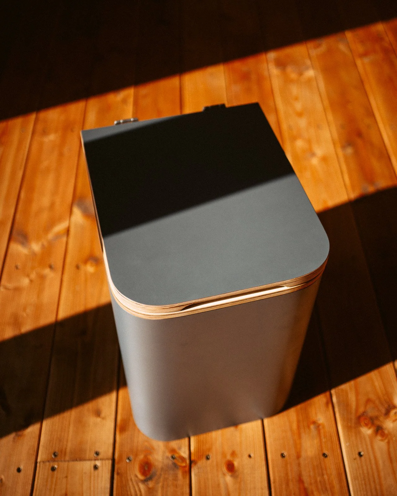 Ein grauer Papierkorb mit einem schwarzen Deckel auf einem Holzboden, durch Sonnenlicht beleuchtet.