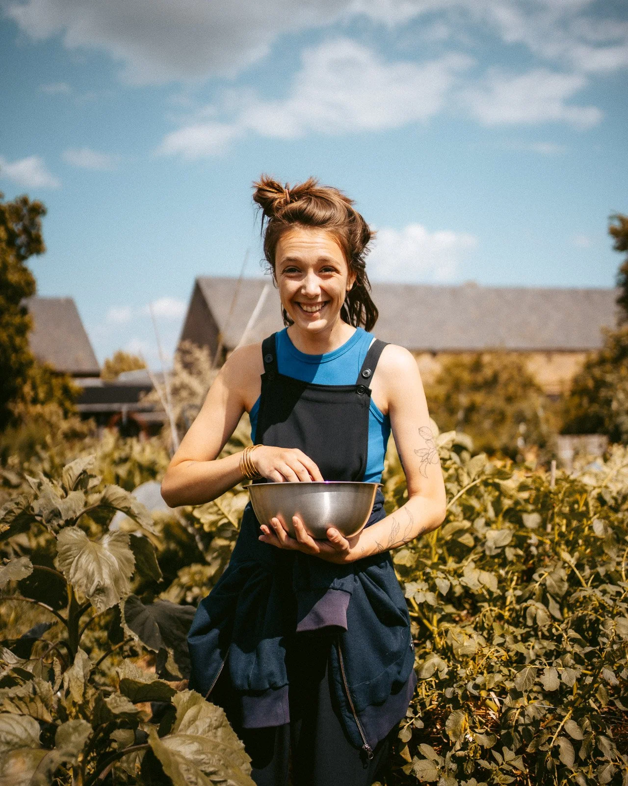 Fröhliche junge Frau im Garten mit blühenden Pflanzen, hält eine Metallschüssel und trägt ein schwarzes Kleid. Im Hintergrund sind Häuser und ein blauer Himmel mit Wolken zu sehen.