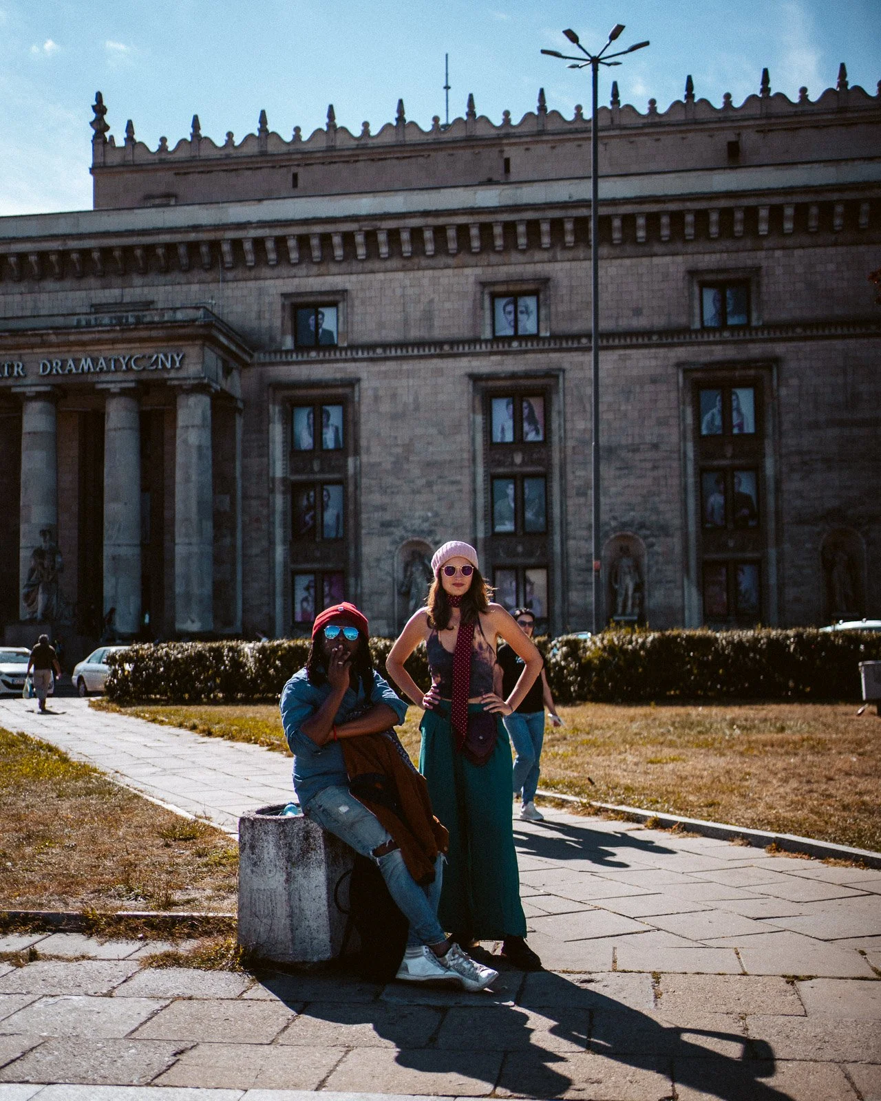 Zwei Frauen mit Sonnenbrillen posieren vor einem historischen Gebäude in der Stadt, im Vordergrund sitzt eine Frau auf einem Betonblock, die andere steht davor.