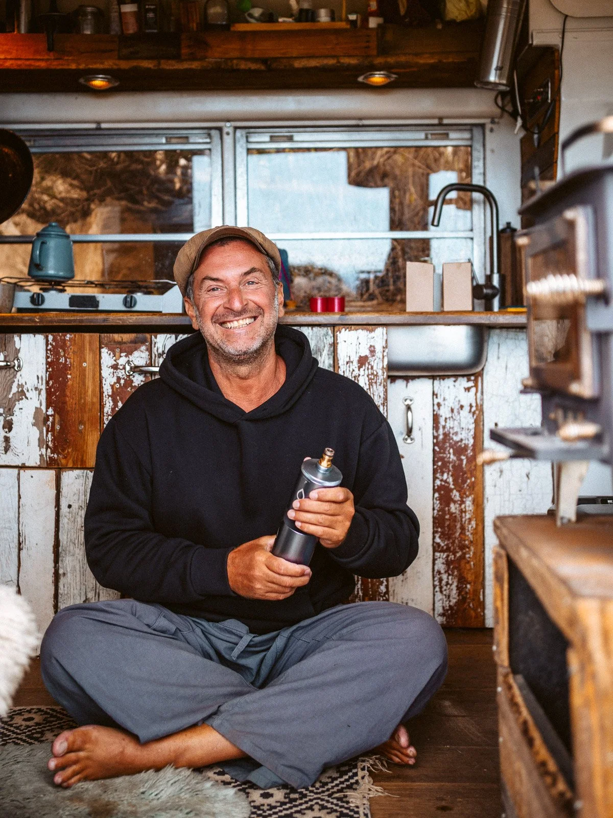 Ein lachender Mann sitzt barfuß unter der Küchenzeile in einer rustikalen Wohnung, hält eine Kaffeemühle und hat einen breiten, freundlichen Ausdruck.