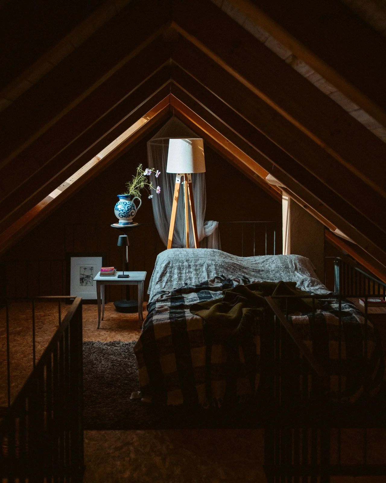 Schlafzimmer im Dachgeschoss mit Holzdecke, Bett, Nachttisch, Stehlampe, Blumentopf mit Blumen und einem Fenster mit Vorhängen.