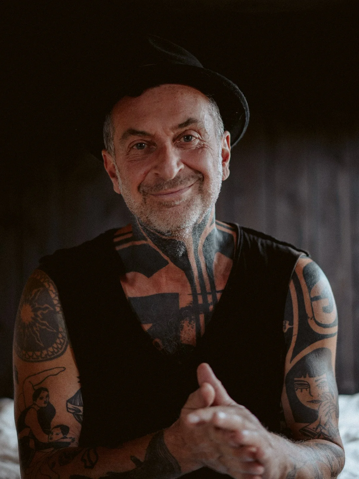 Ein lächelnder Mann mit Tattoos auf Armen, Schultern und Hals trägt einen schwarzen Hut und ein schwarzes T-shirt, sitzt vor dunklem Hintergrund.