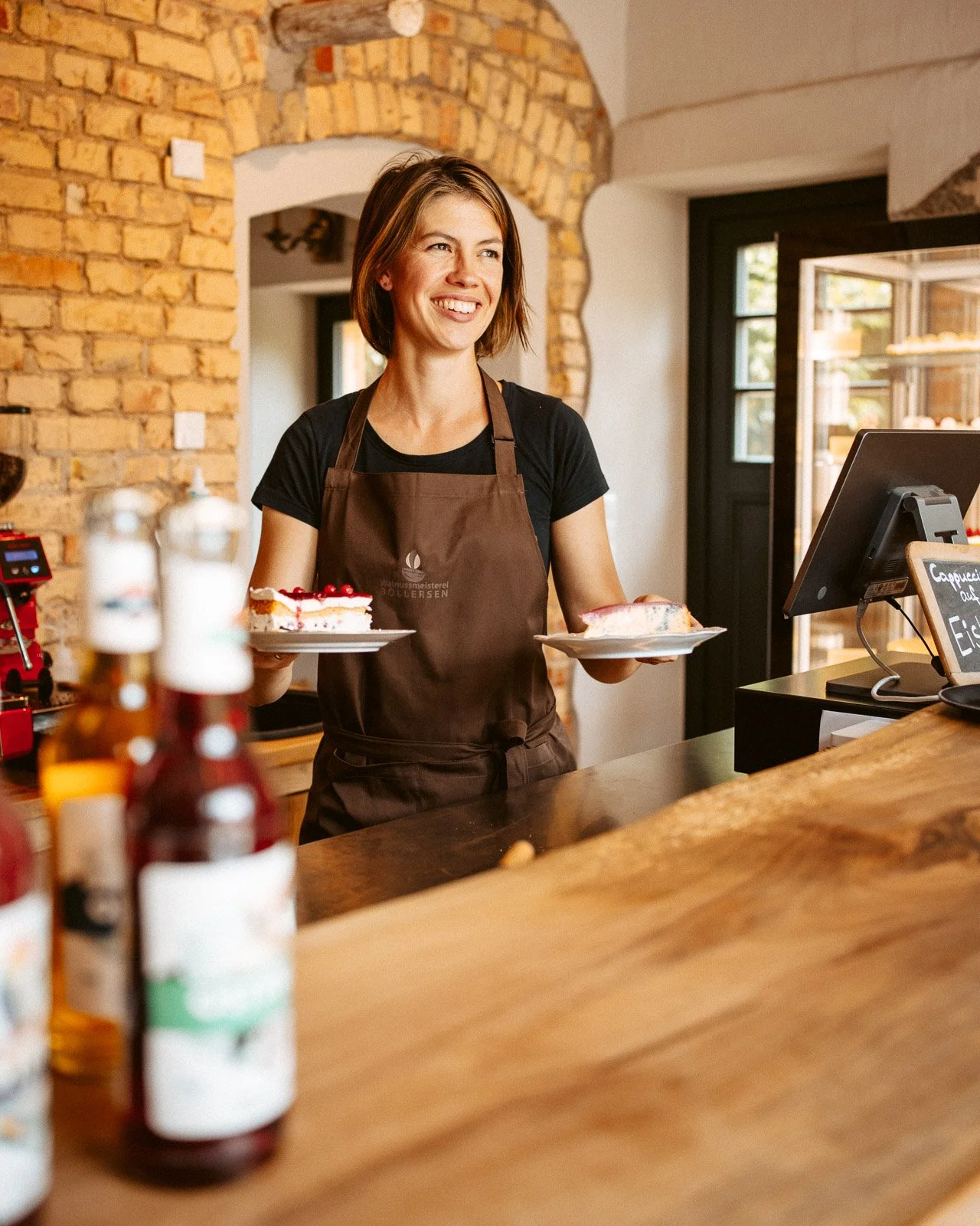 Eine lächelnde Frau mit braunem Haarschneidet im schwarzen T-Shirt und brauner Schürze steht hinter einer Theke und hält zwei Kuchenplatten mit Kuchen, im Hintergrund eine Steinausgangswand mit einem Türrahmen und einem Fenster, im Vordergrund sind e