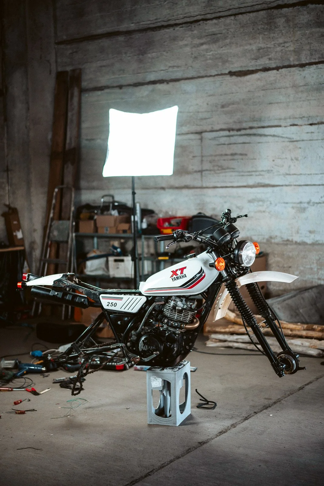 Ein Yamaha XT 250 Motorrad in einer Werkstatt, auf einem Ziegelstein gesetzt, um den Motor zu stützen, mit Werkzeugen und Werkstattmaterialien im Hintergrund.