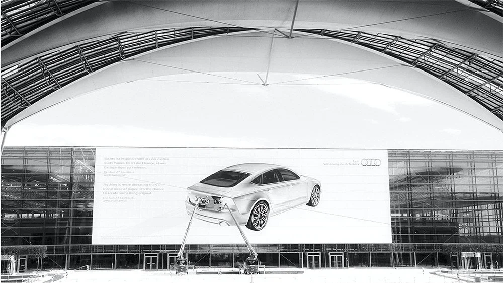 Ein großes Plakat eines weißen Autos der Marke Audi, das an einer Glasfassade eines Gebäudes angebracht ist, mit einem modernen, architektonisch auffälligen Dach.